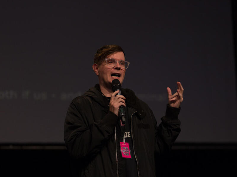 A person with short hair and glasses speaking into a microphone. Their left hand is holding the microphone and their right hand is up in the air gesturing. 