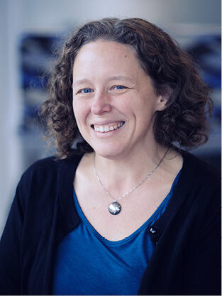 A portrait of a woman with short brown hair wearing a blue shirt, black cardigan, and a silver necklace with a circle shaped pendant. 