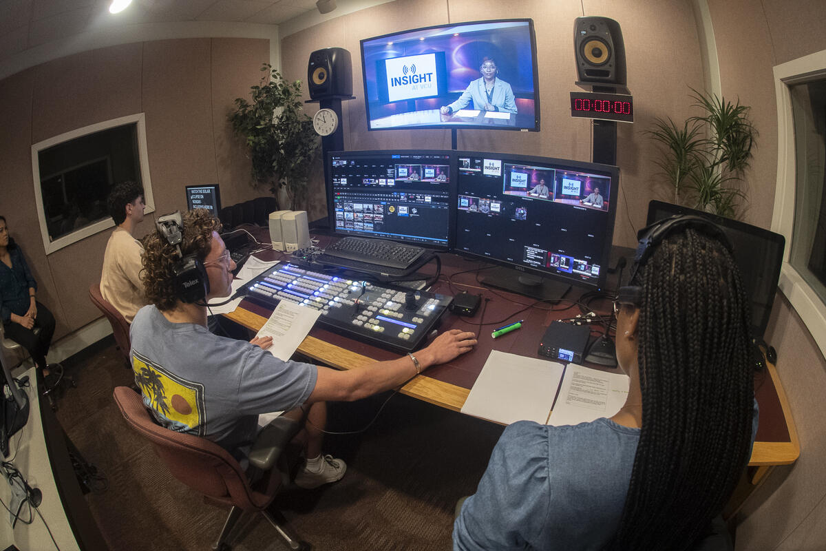 A photo of two ment sitting at a seks with comptuers and sound editng eiquiment on it. A woman near them is standing to the right, and another is sitting in a chair on the left. 