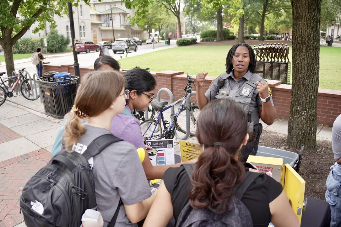 A photo of a police officer talking to four people at a table outside. 