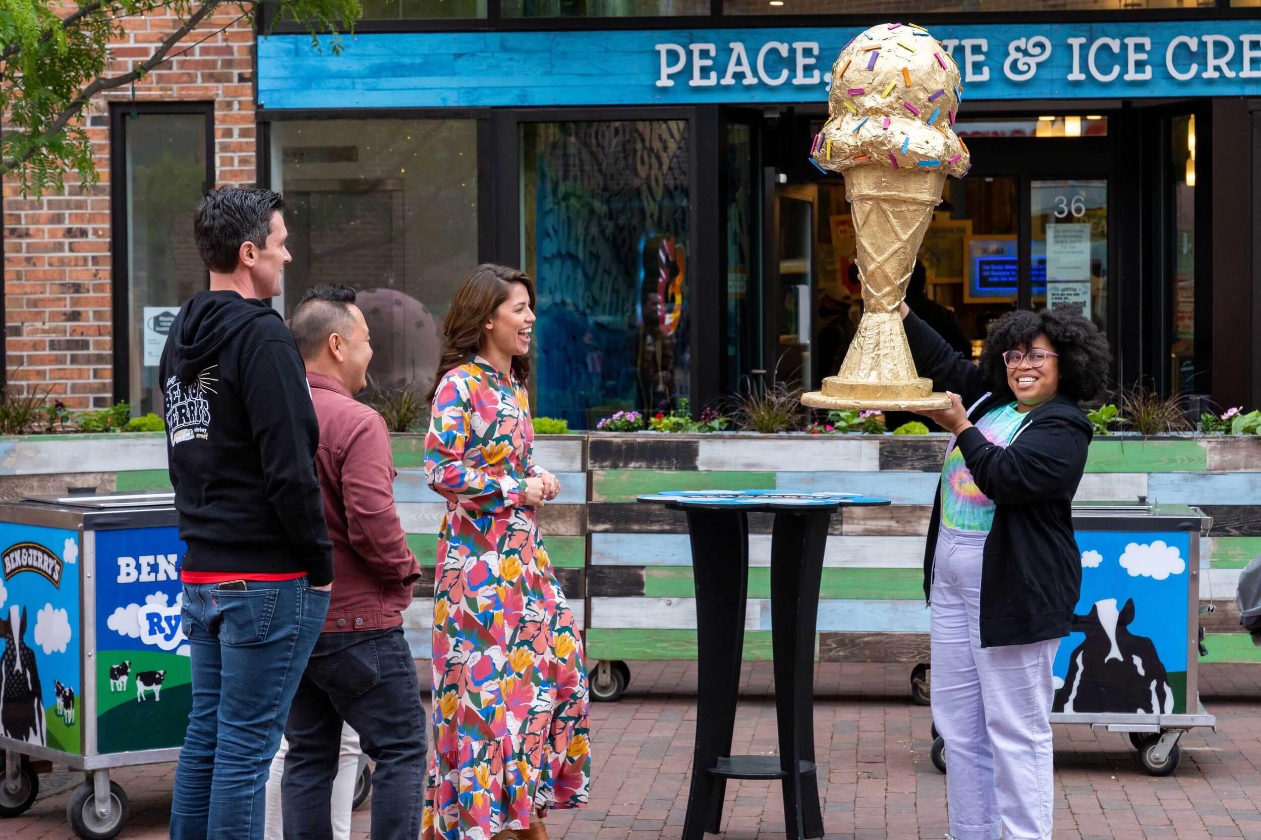 A woman holds aloft a large ice cream cone trophy. Two men and a woman stand to the side, watching her with smiles.