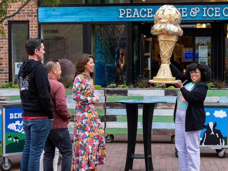 A woman holds aloft a large ice cream cone trophy. Two men and a woman stand to the side, watching her with smiles.