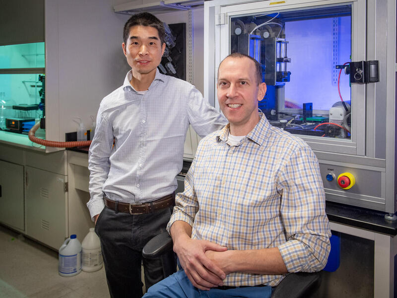 A photo of a man standing to the left of a man sitting in a chair. 