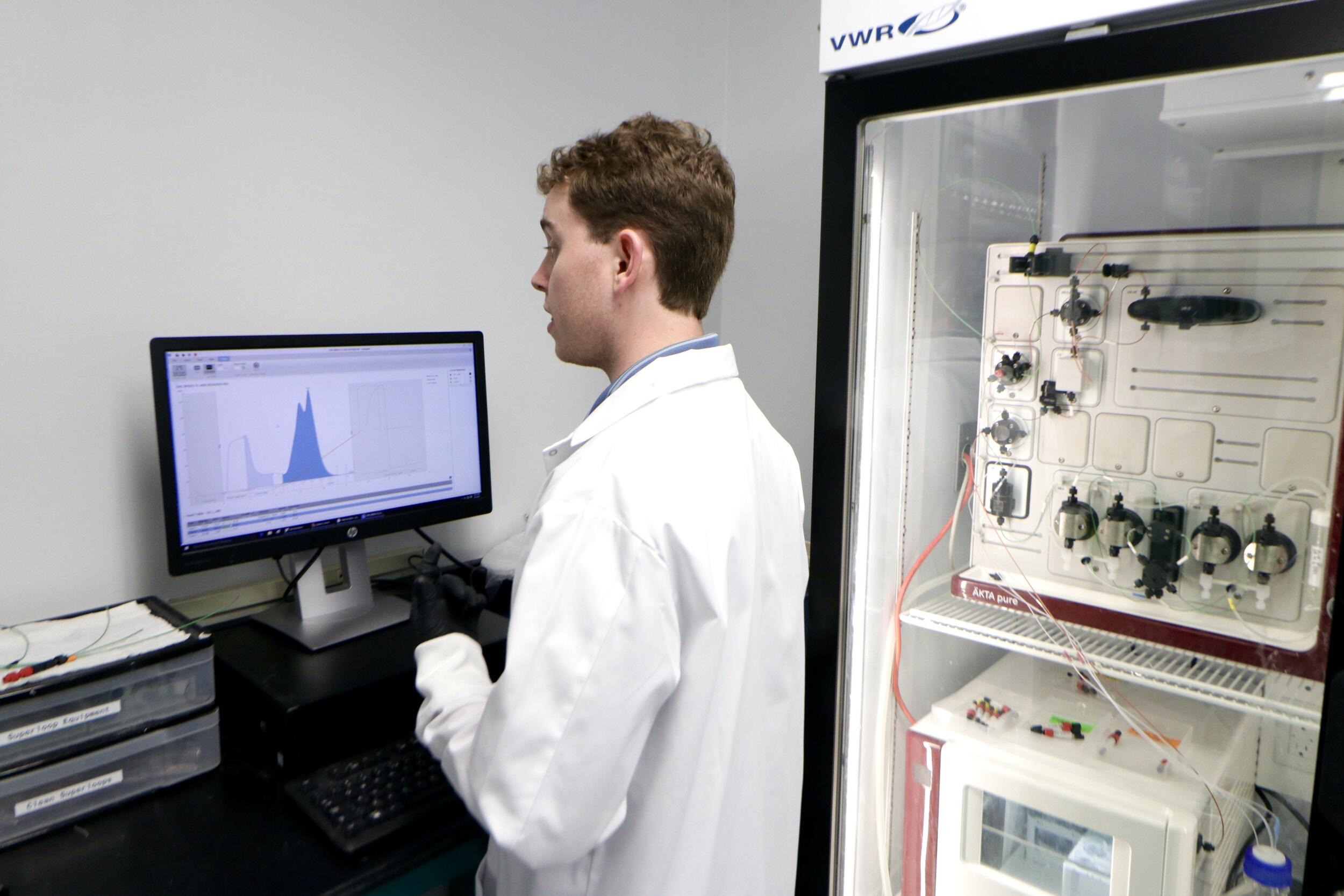 A photo of a a man wearing a white lab coat standing in front of a computer monitor. 