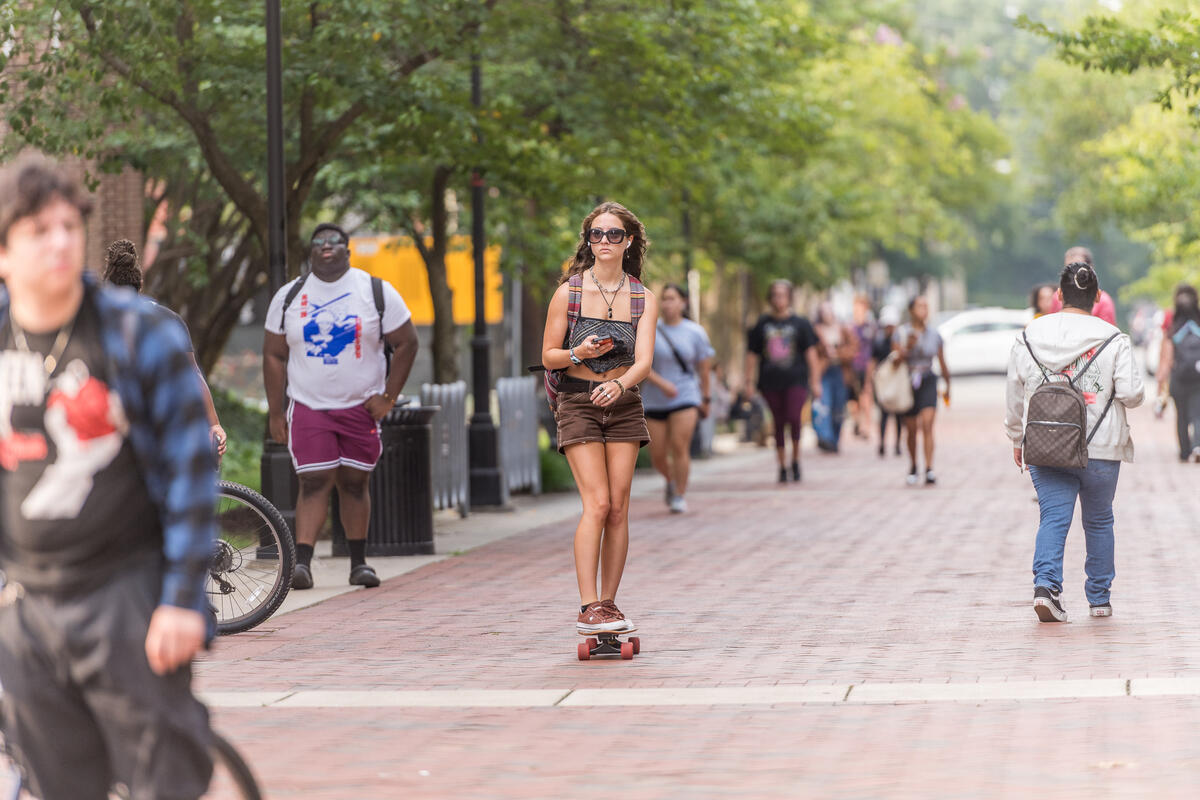 A photo of a woman skateboarding down a sidewalk while people walk around her. 