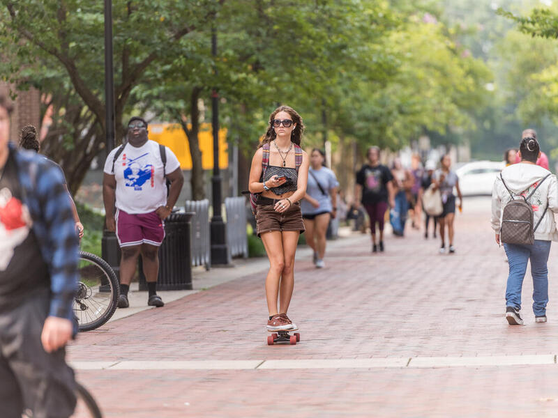 A photo of a woman skateboarding down a sidewalk while people walk around her. 
