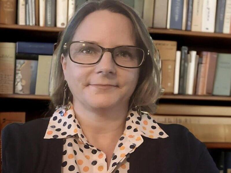 A portrait of a woman from the chest up in front of a book case. 