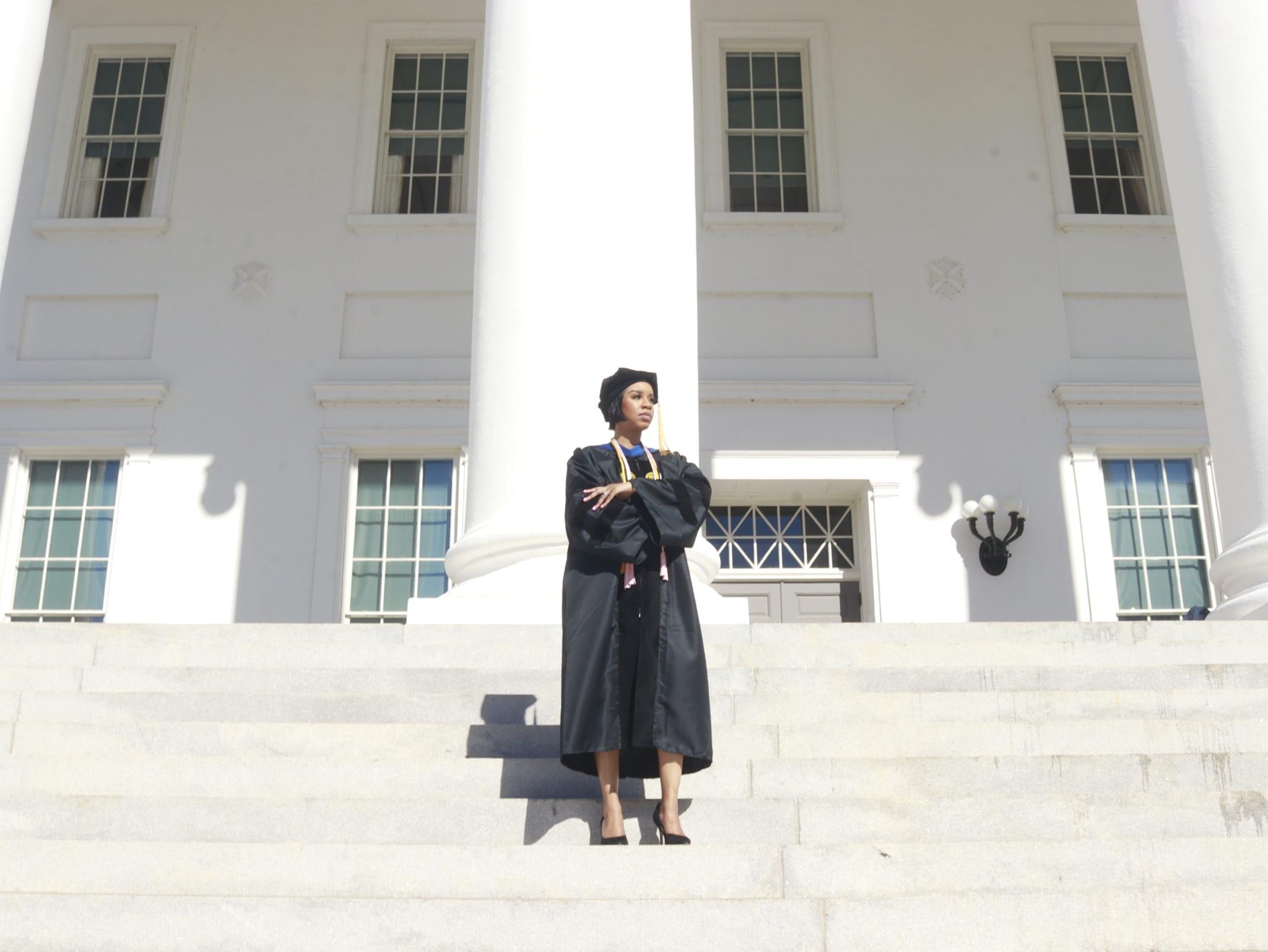 A photo of a woman in a doctoral graduation cap and gown standing on white steps in front of a white building. 