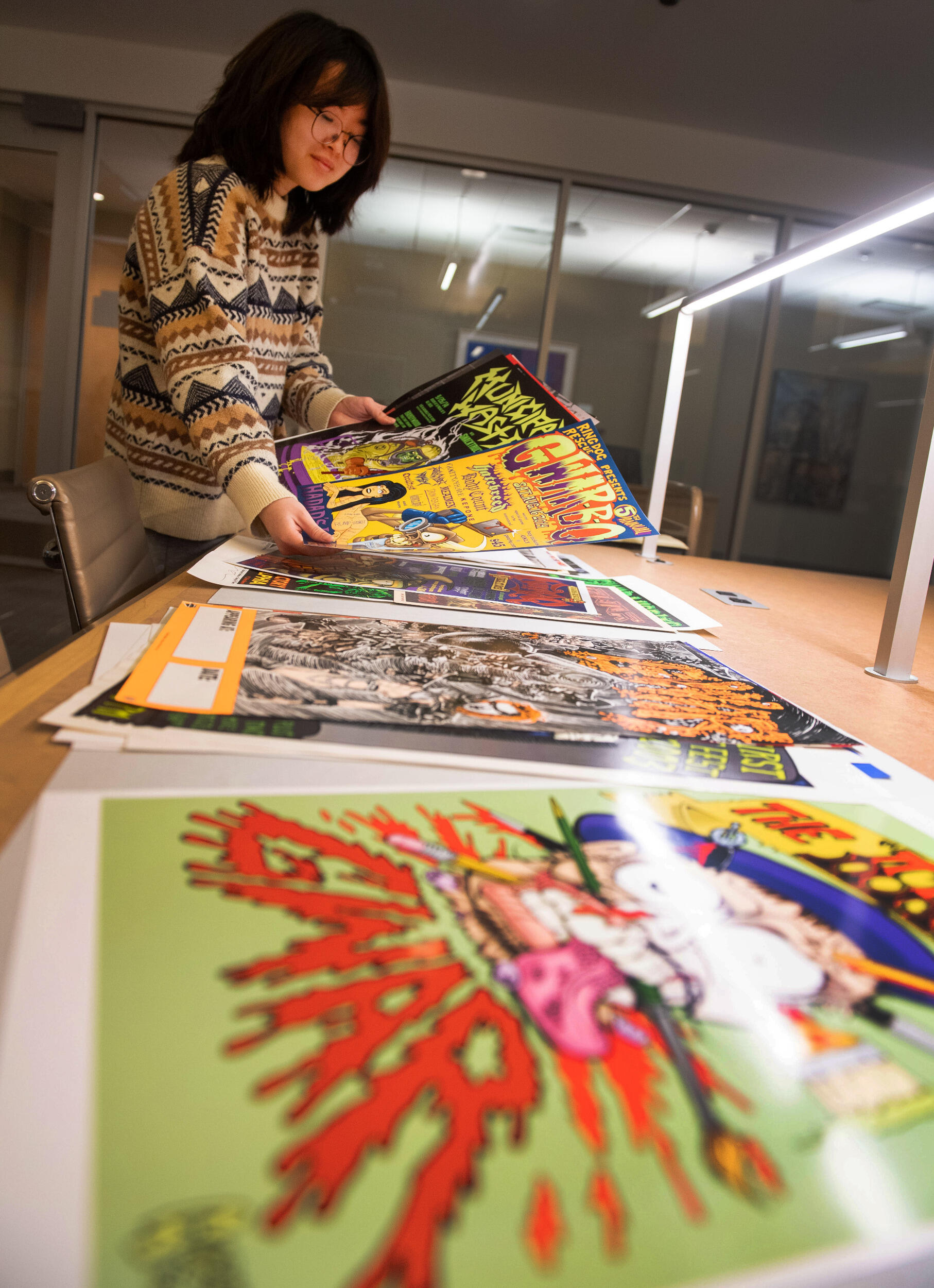 A person in a sweater and glasses looks at posters arrayed on a table in front of them.
