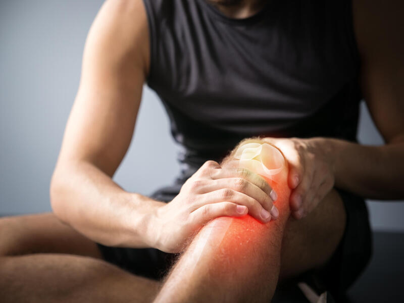 A photo of a man from the chest down sitting on the ground. One he has his hands resting on one knee, which has been illustrated over, so you can see the knee bone. 