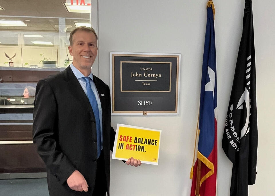 A photo of a mant from the waist up. He is holding a yellow sign that says \"SAFE BALANCE IN ACTION.\"