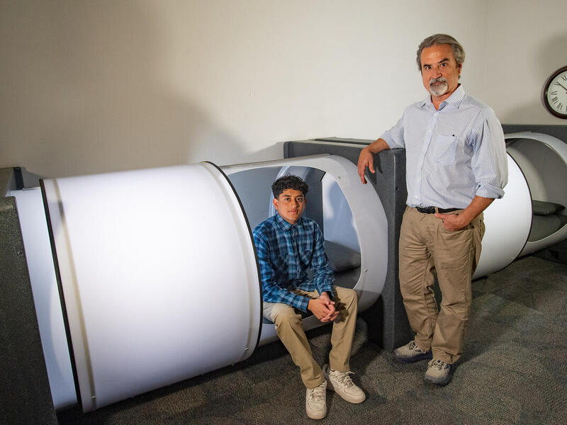 A photo of a man sittin in a white pod, and another man standing to the right of the pod, resting his left arm on the side of it. 