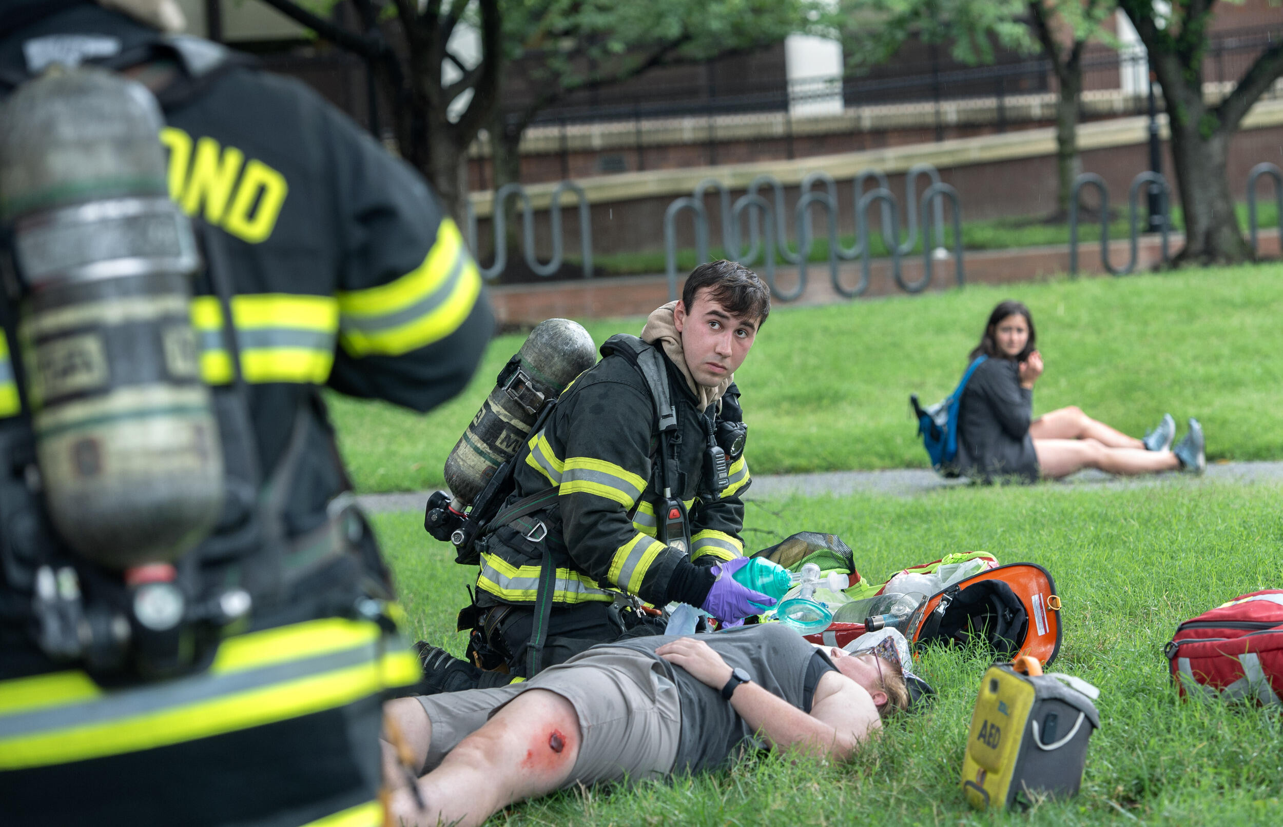 A photo of a firefighter using an oxegen mask on a person laying on the ground. The person laying has a bloody wound on their leg. In the background, another person sitting is looking at the firefighter and person laying. In the foreground is the back of a firefighter. 