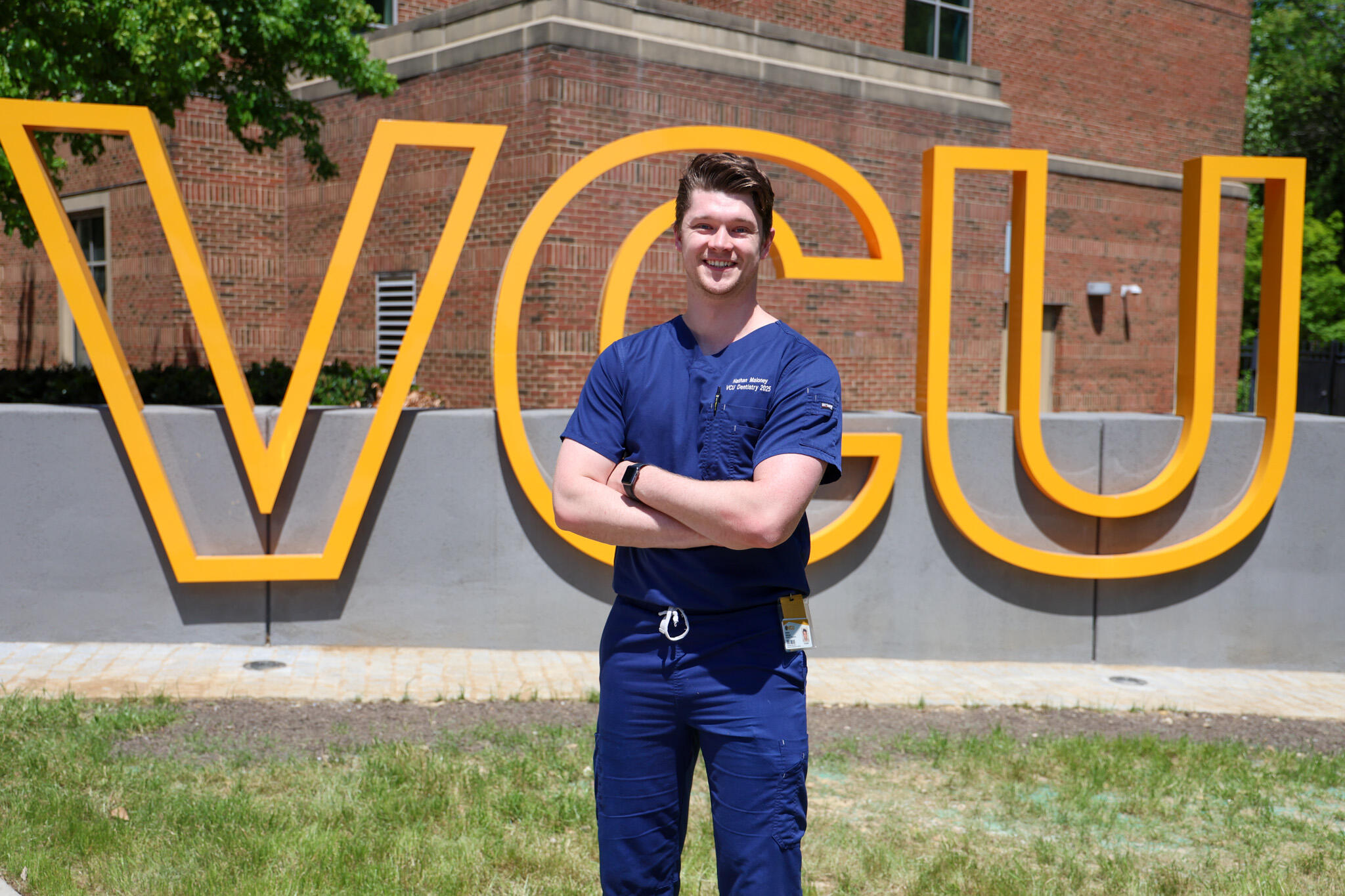A photo of a man wearing blue dental scrubs from the knees up. He has his arms crossed against his chest and is standing in front of a sign that says \"VCU\" in large yellow letters. 