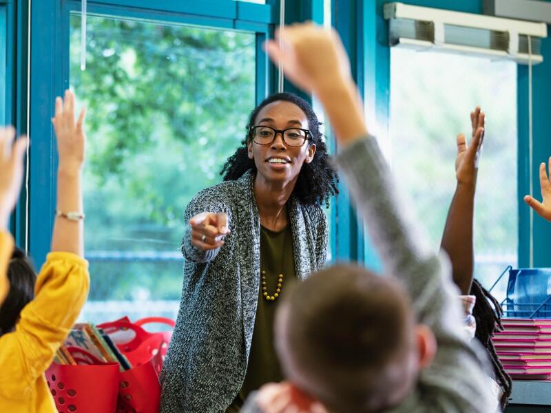 A photo of a teacher standing in front of a row of students who are raising their hands