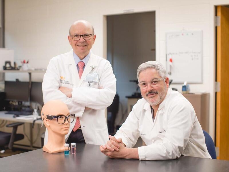 One doctor standing with his arms crossed across his chest and another sitting down. There is a table in front of them with a mannequin head wearing black rimmed glasses. 