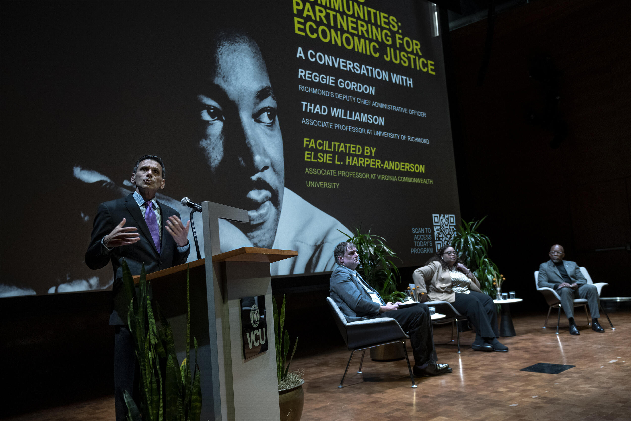 A man standing at a podium speaking. To his right, three people sit in chairs on a stage. 