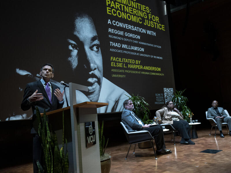 A man standing at a podium speaking. To his right, three people sit in chairs on a stage. 