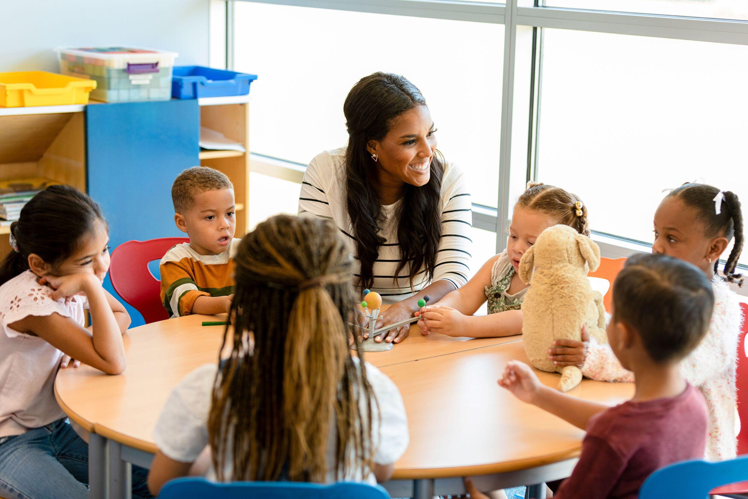A teacher sitting at a round table with five young children. 