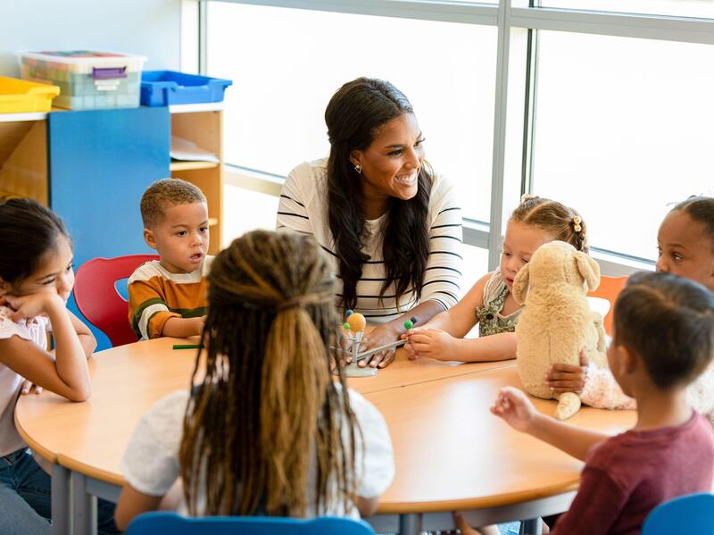 A teacher sitting at a round table with five young children. 