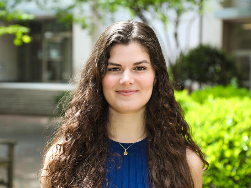 A photo of a woman from the shoulders up standing outside. She is standing in front of bushes and the entrance to a building. 