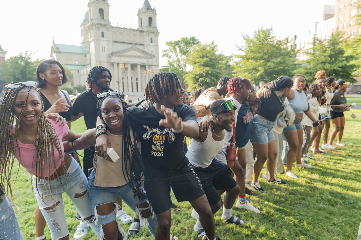 A photo of a group of students linking eachother into a long chain by putting their arms around the person whose next to them's shoulders or waist. 