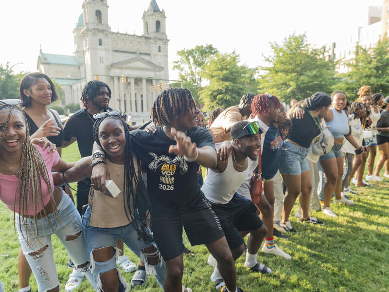 A photo of a group of students linking eachother into a long chain by putting their arms around the person whose next to them's shoulders or waist. 