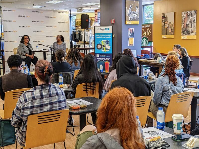 Two women sitting in front of a crowd of people. The woman on the left is speaking 