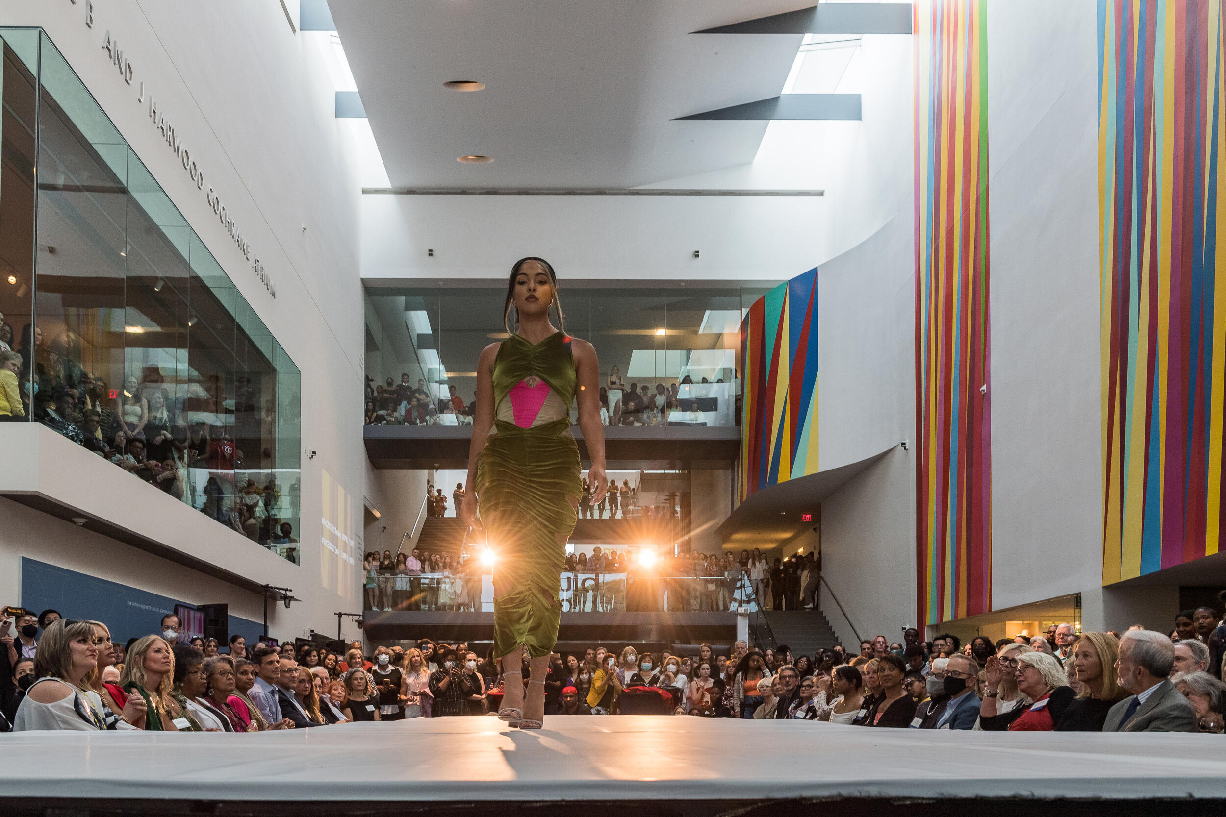 A model walks along a runway in a large brightly-lit atrium lined with art on the walls as an audience watches on either side of the runway.
