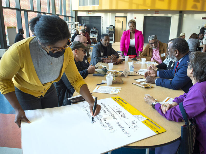 A photo of a woman writing on a giant pad of paper. Behind her six more people are sitting and standing around the table. 