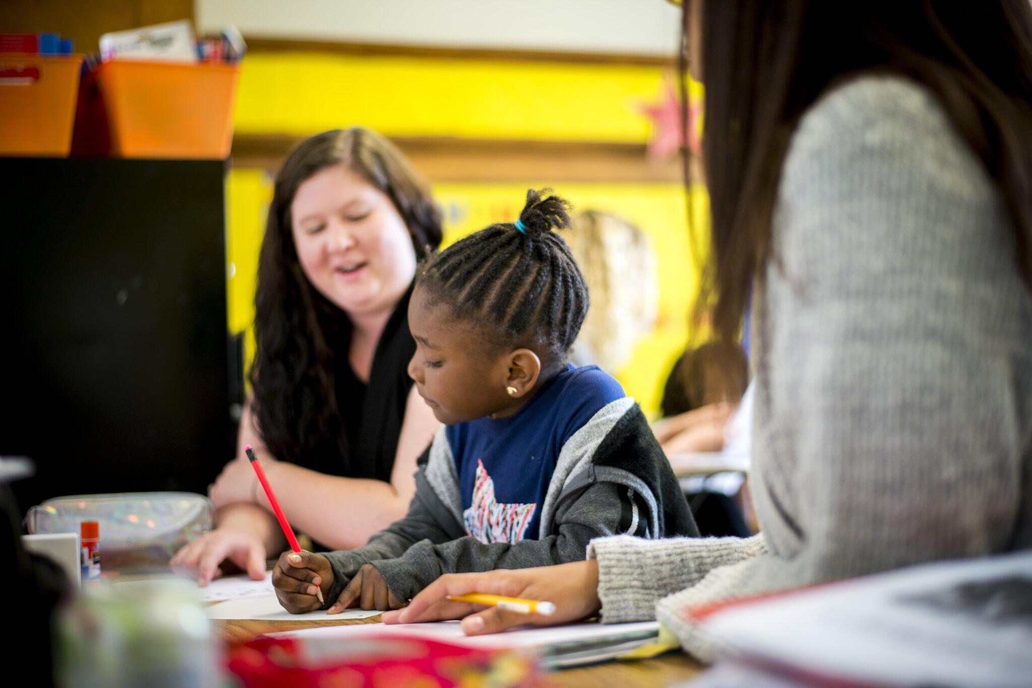 A woman sitting with a girl at a table while she works on a work sheet 