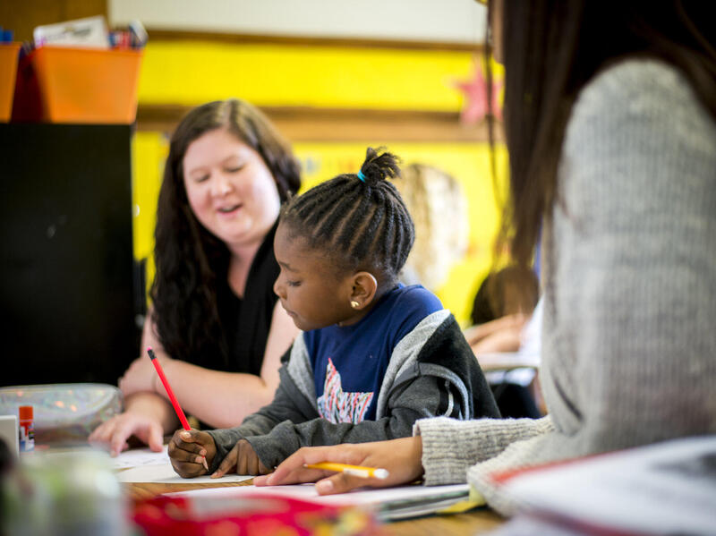 A woman sitting with a girl at a table while she works on a work sheet 