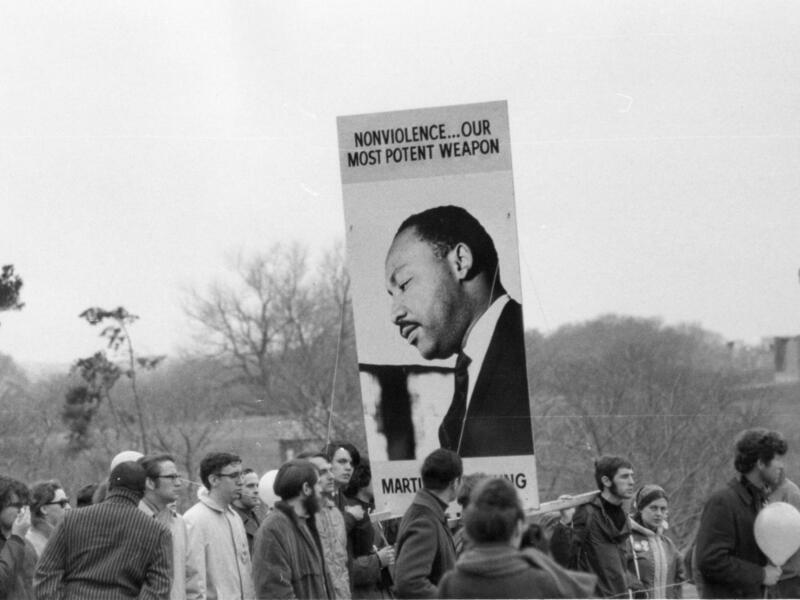A black and whie photo of a crowd of people. In the middle of the crowd is a sign with a photo of Martin Luther King Jr. that says \"NONVIOLENCE.... OUR MOSTPOTENT WEAPON\" 