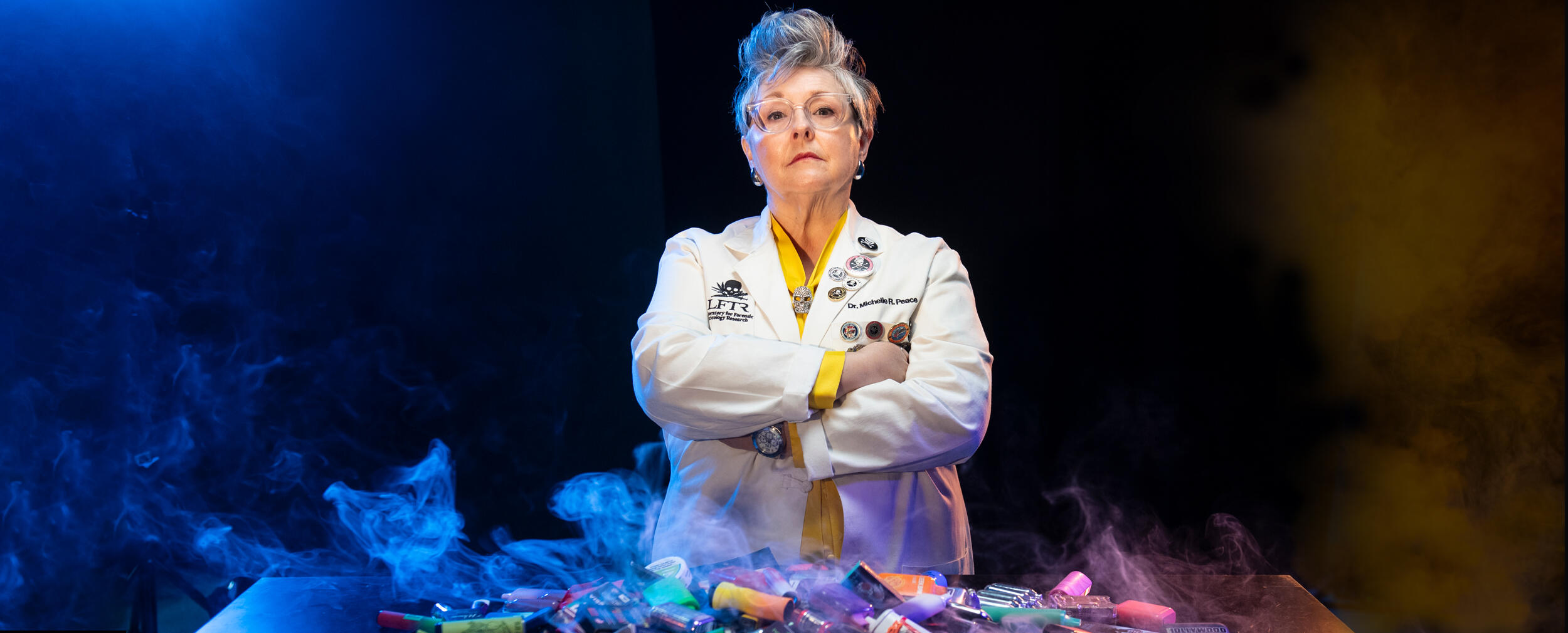 Professor Michelle Peace wears a lab coat and stands in front of a table with vaping paraphernalia and colorful smoke.
