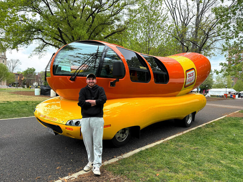 A photo of a man standing in front of a car that is shaped like a hot dog. 