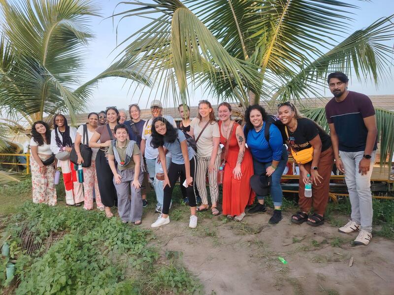 A group photo of 16 people bending over in front of some short palm trees. 