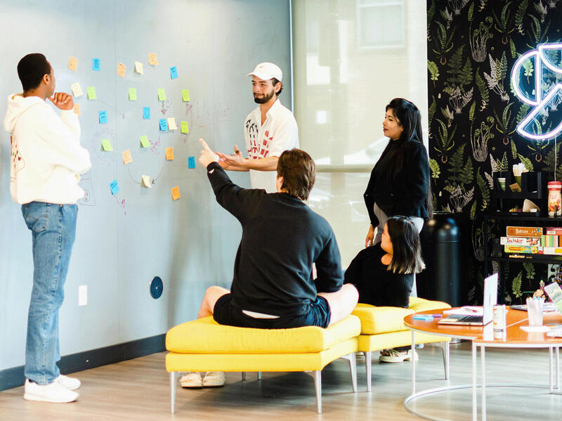 A photo of three people standing and one person sitting in front of a wall with sticky notes on it. 