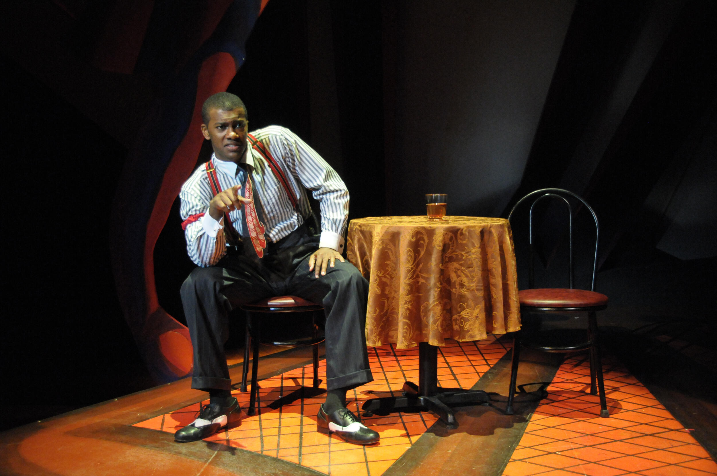 An actor on a stage, sits on a chair in the spotlight and speaks.