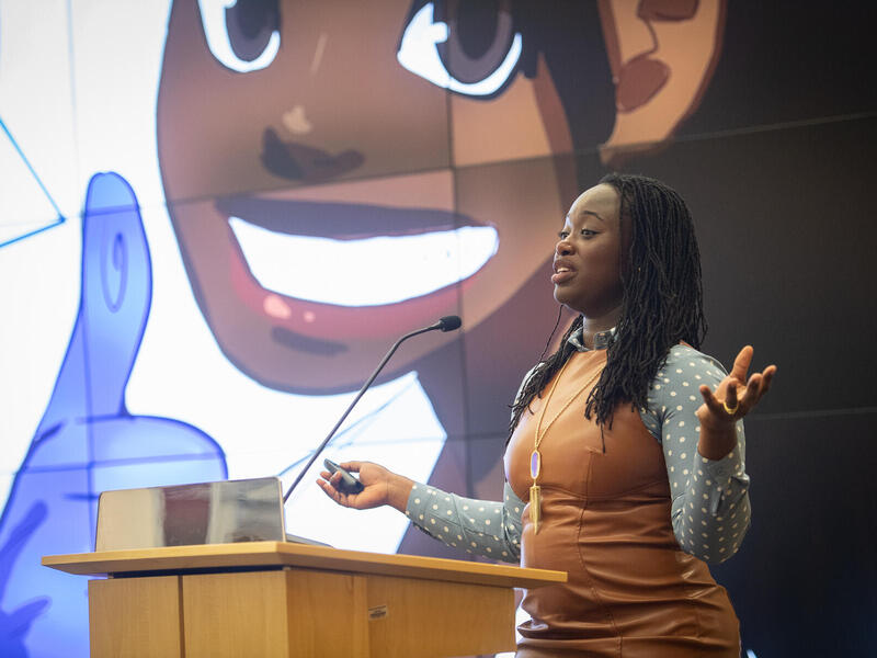A photo of a woman speaking while standing behind a podium. Behind her is a projection of an illustration of herself. 