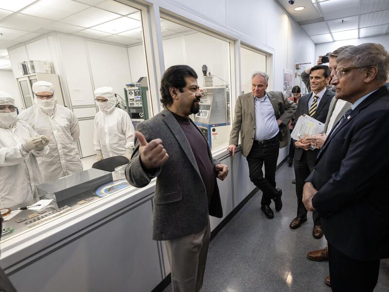 A man talking to a group of people in front of glass windows. On the other side of the windows people in white protective gear are in a lab 
