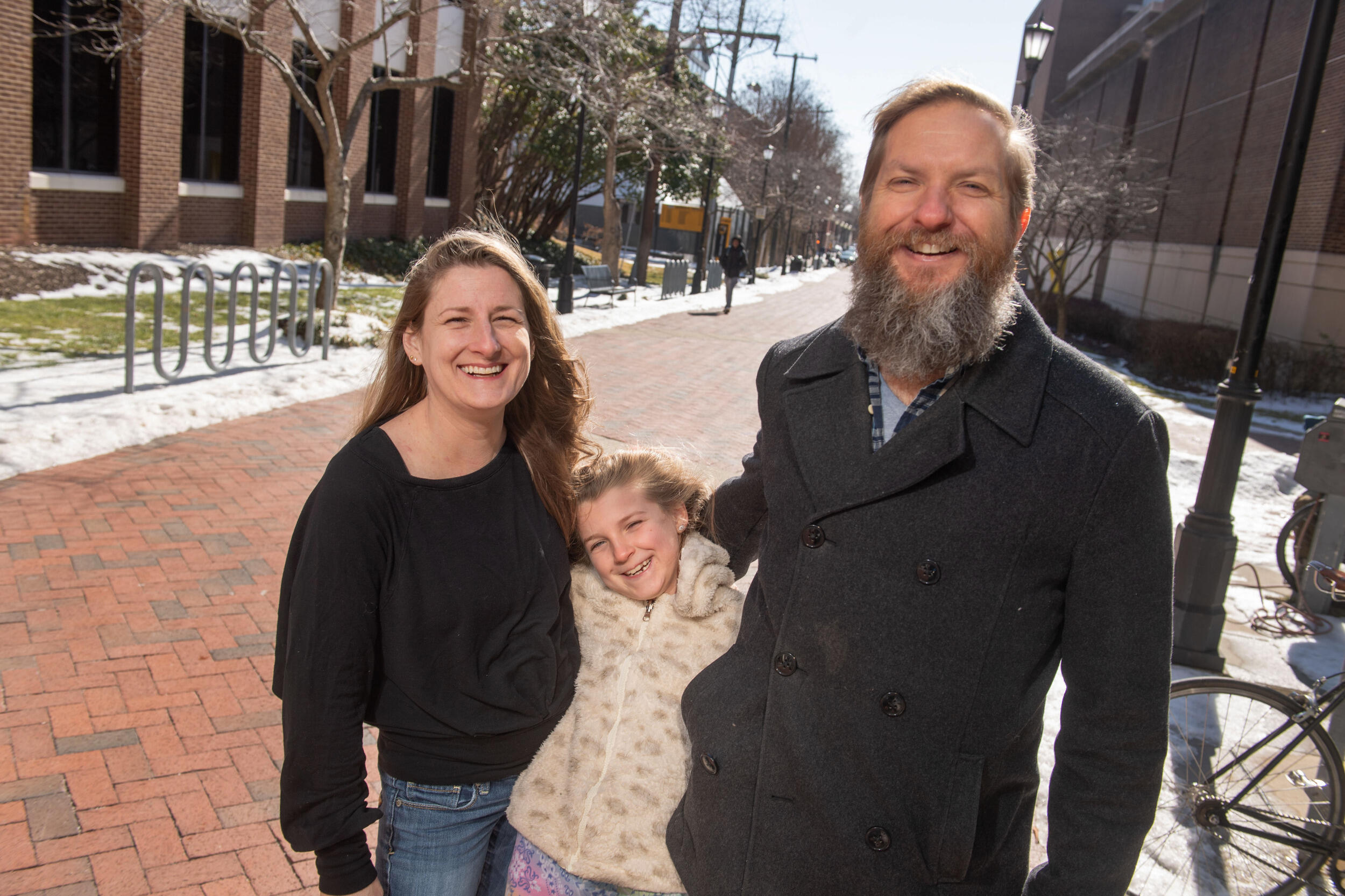 A woman and man stand on a long walkway with a young girl between them. All are smiling.