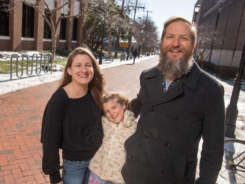 A woman and man stand on a long walkway with a young girl between them. All are smiling.