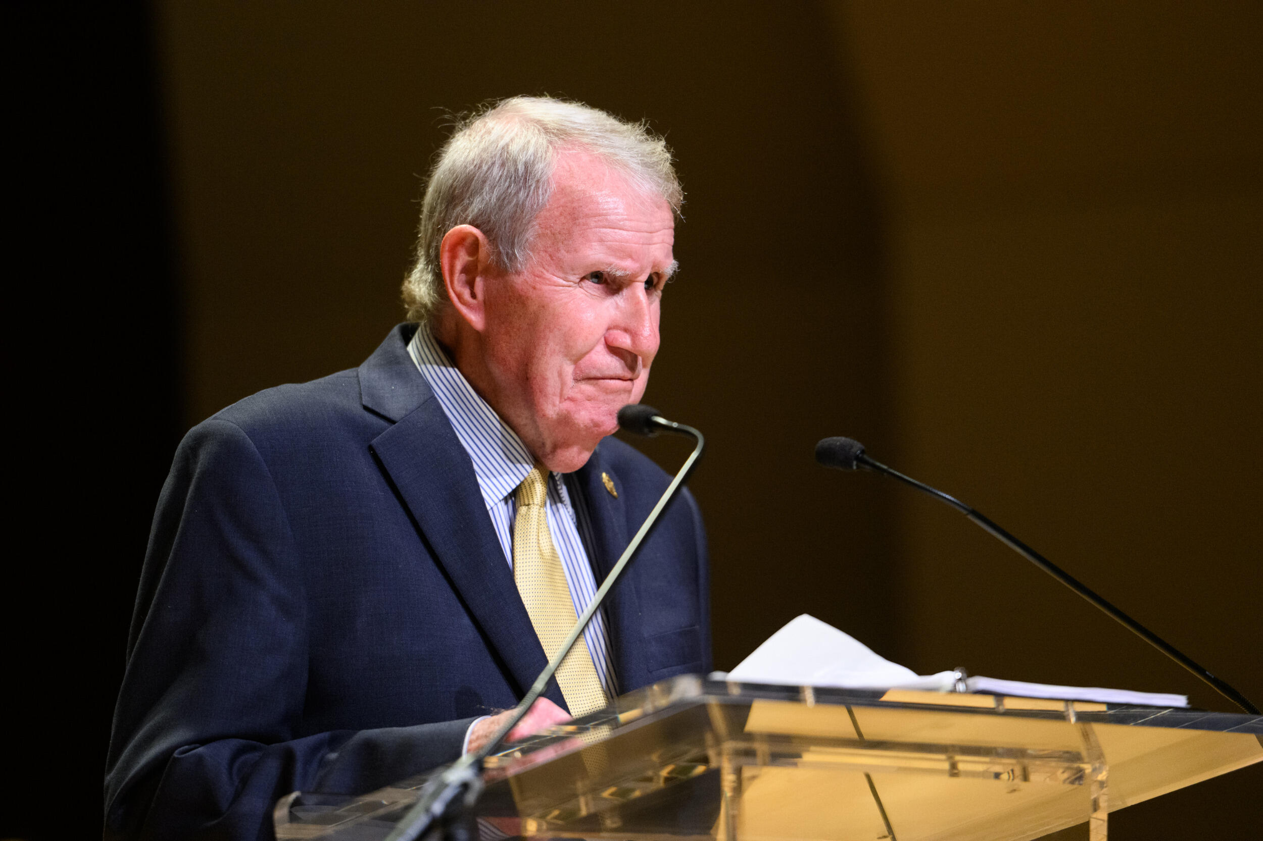 A man in a coat and tie speaks into a microphone.