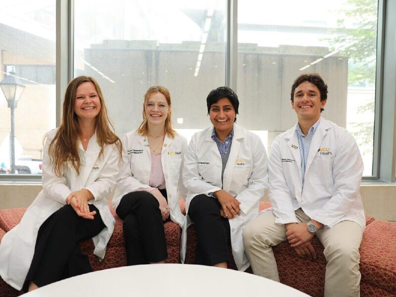 A photo of four people sitting on orange plush benches aligned in a semi-circle. Everyone is wearing a white doctor's coat.