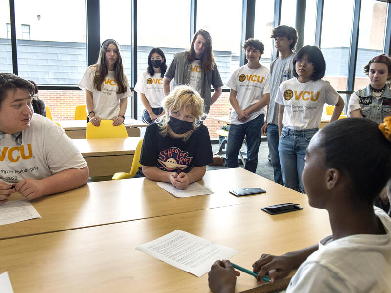 A photo of three people sitting at a table and a croup of people standing behind them. 