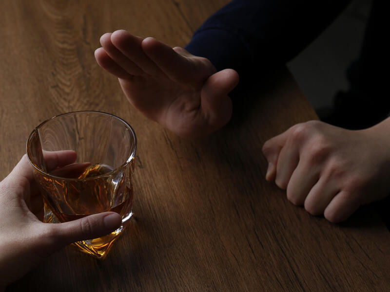 A hand on the left is holding a cup with brown liquid and a hand on the right is pointing upward in a \"stop\" gesture. 