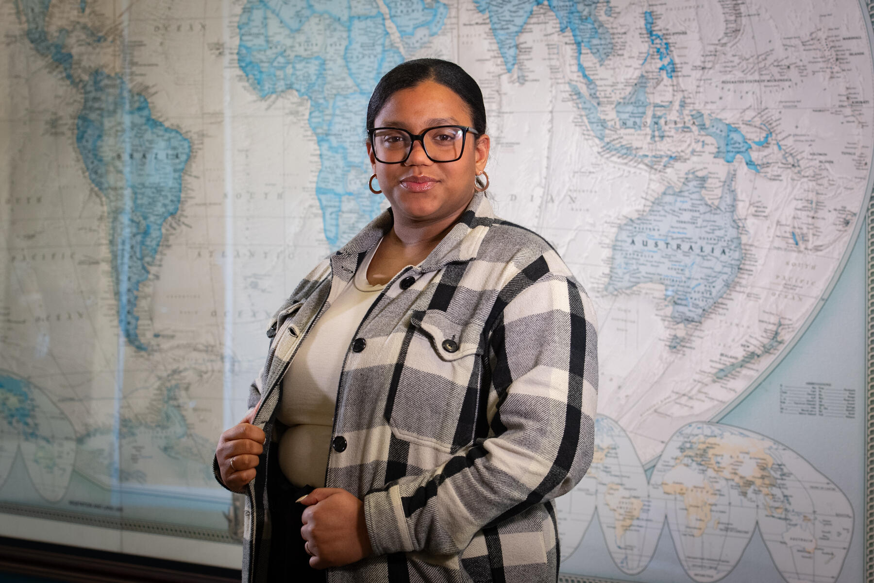 A photo of a woman from the waist up. She is standing in front of a map of the world. 