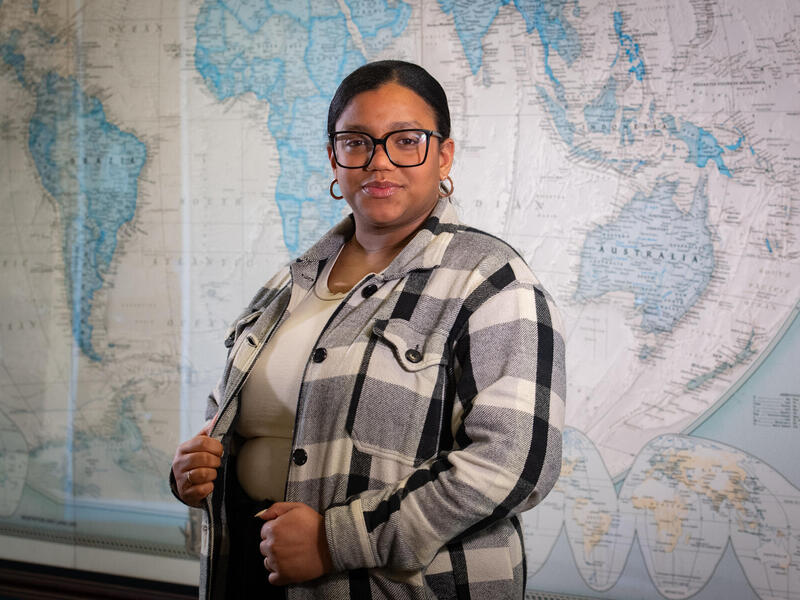 A photo of a woman from the waist up. She is standing in front of a map of the world. 