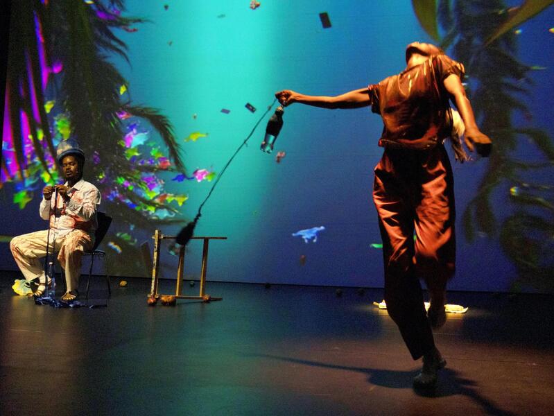 A photo of two people on a stage. The person on the left is sitting on a stool, and the person on the right is dancing. 