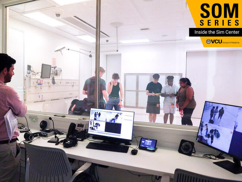 A photo of a man standing behind a desk looking through a glass window. He is watching five people interacting on the other side. 